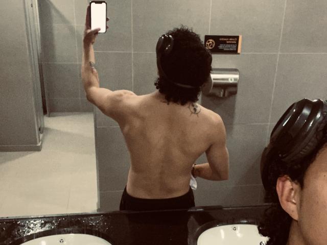 Jay Taylor, ohne Hemd, mit lockigem, schwarzem Haar, muskulösem Rücken und einem Tattoo auf dem Arm, macht ein Selfie im Badezimmer und zeigt sein nacktes Gesäß in die Kamera..