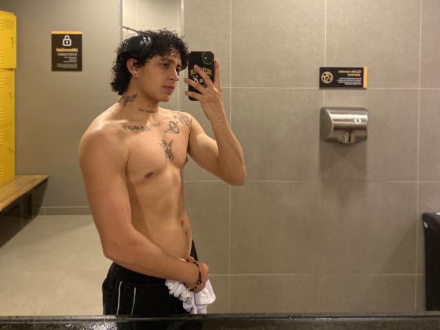 JayTaylor sans chemise, avec des cheveux noirs bouclés, des tatouages sur la poitrine et le cou, tenant un téléphone pour un selfie devant un miroir, pantalon noir, serviette blanche, une main couvrant ses fesses, prenant une photo dans la salle de bain..