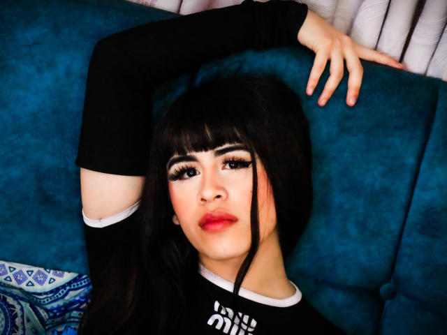 HannaMitsury with black hair, heavy eyeliner, and red lipstick, arms raised, lying on blue couch, wearing black long-sleeve top, looking at camera.