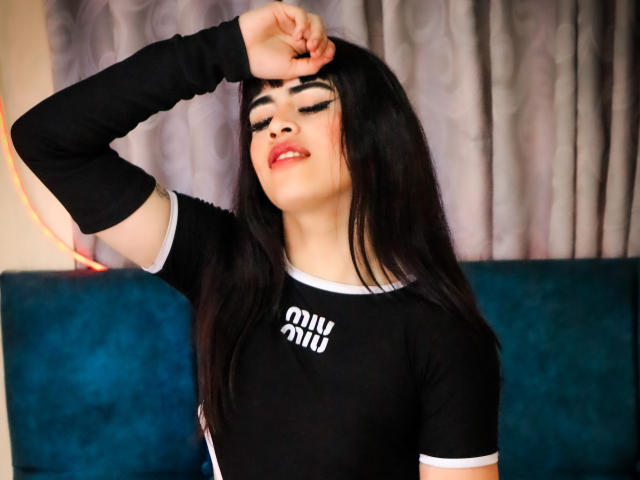 HannaMitsury with black hair, eyes closed, smiling, arm raised, wearing black long-sleeve shirt, white trim, and bold makeup, standing in front of blue velvet couch.