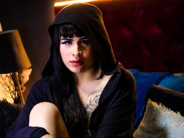 HannaMitsury with black hood, long straight hair, dark eyeliner, tattoo on chest, black hoodie, sitting, leg up, lamp on left, warm light, blue pillow behind, soft expression.