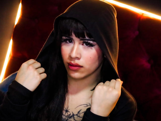 HannaMitsury with long black hair, wearing a black hoodie, hands on hood, has a tattoo on her chest, dark makeup, and pink lipstick, with a serious expression.