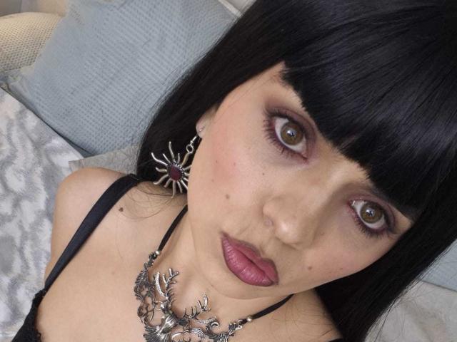 CatalinaRuiiz has pale skin, brown eyes, black bangs, and wears a black necklace with leaf designs, purple lipstick, and spider earrings. Her tits are small and she's looking up.