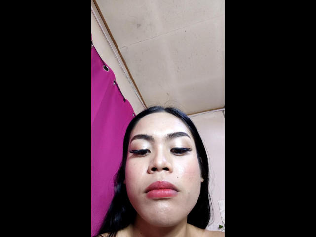 AryanaGwafa with shiny, silver eyeshadow, thick black eyebrows, and pink lips, looking down at the camera with a neutral expression, black hair, and a slightly sweaty forehead.