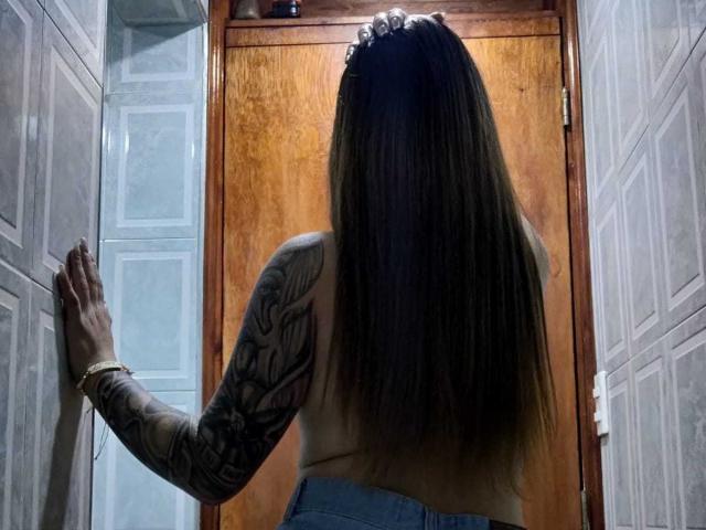 Topless Catag with long brown hair, tattooed arm, and blue jeans stands against a wooden door, hand on wall, back to camera, wearing a bracelet.