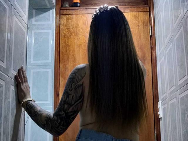 Tattooed Catag with long brown hair, back to camera, wearing a tank top and blue jeans, stands with hands on white doors, wooden door in front.