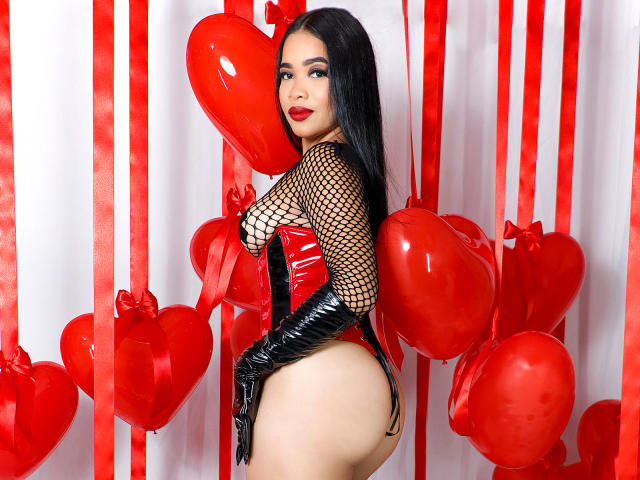KarlaBella with long black hair, wearing a red and black fishnet top, black gloves, and a red latex bodysuit, showing her big ass and small thong, surrounded by red heart balloons.