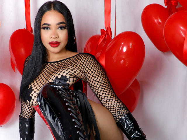 KarlaBella with long black hair, red lipstick, fishnet top, black leather corset, and glossy black boots, sitting among red heart balloons, showing her shiny black boots and thighs.