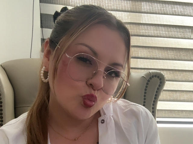 LaurenSpitz with puffy red lips, making a pouty face, wearing large round glasses, white shirt, gold hoop earrings, and a small black hair bow, in a chair.