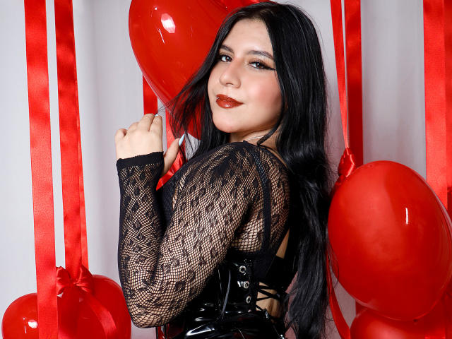 DannaOlivieri with long black hair, wearing a black mesh top, holding red balloons, looking over her shoulder, smiling with red lipstick, showing her ass in a black corset.