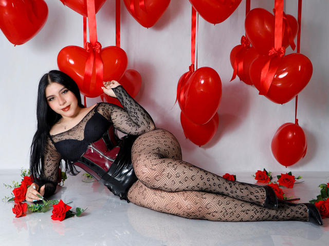 DannaOlivieri with black lace bodysuit, red corset, and fishnet legs, lying on floor with red roses, surrounded by red heart balloons, and black high heels.