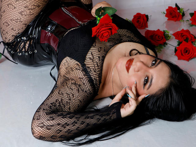 DannaOlivieri, with black fishnet top, red roses, and black leather shorts, laying down, holding rose, red lipstick, long black hair, sexy, curvy, looking at camera, soft smile.