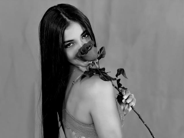 AvaMorgan with long dark hair, holding a rose to her nose, looking over her shoulder, wearing a lacy dress, in a black-and-white photo. Boobs, rose, AvaMorgan's face.