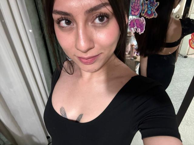 Luluberrie with medium breasts, black top, and tattoos, has dark brown hair, brown eyes, and a slight smile, reflected in a mirror showing her back and black bra.