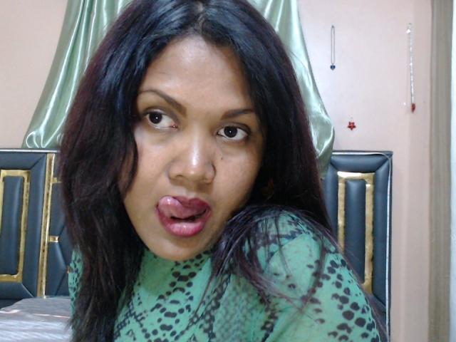 FelyLove has dark, wavy hair, brown eyes, and is sticking out her pink tongue. She's wearing a green, patterned top and looking directly at the camera.