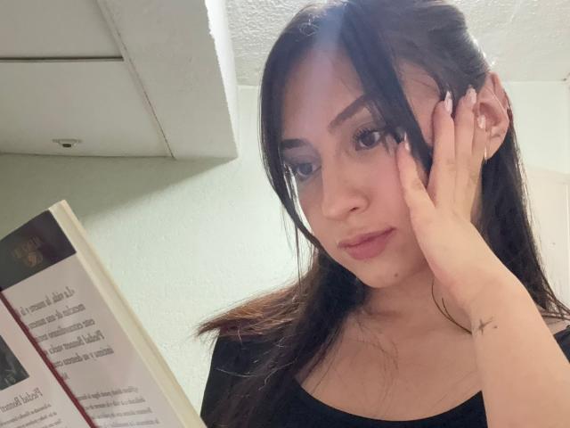 Luluberrie with dark brown hair, black top, and fair skin, holding a book, touching her ear, looking at the camera with a slight smile. Her tits are small.