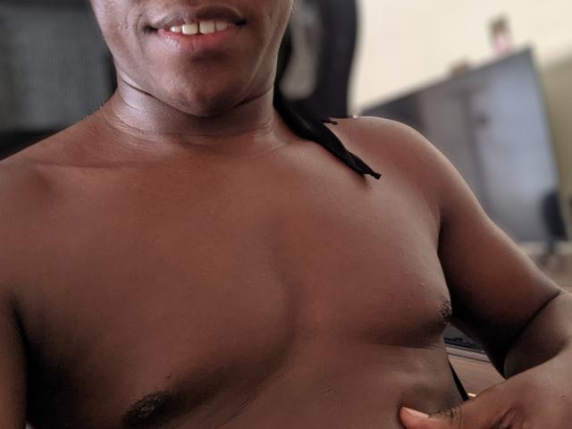 SexyOccy has a dark brown, muscular chest with small, dark nipples and a black strap on their right shoulder. They're smiling, showing white teeth.