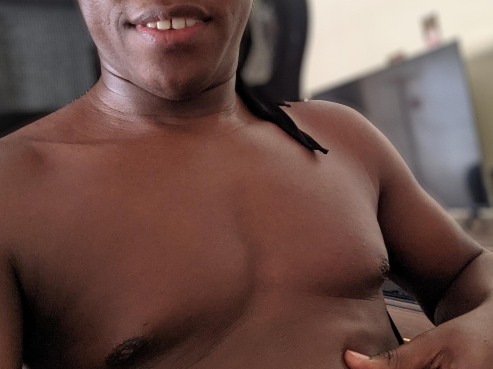 SexyOccy has a dark brown, muscular chest with small, dark nipples and a black strap on their right shoulder. They're smiling, showing white teeth.