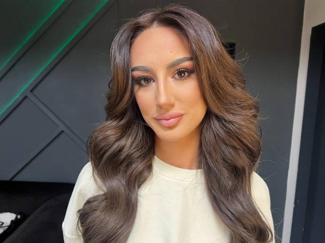 MackenzieHart has long, wavy brown hair, full eyebrows, and is wearing a white shirt. Her lips are slightly puckered in a subtle, sexy smile.
