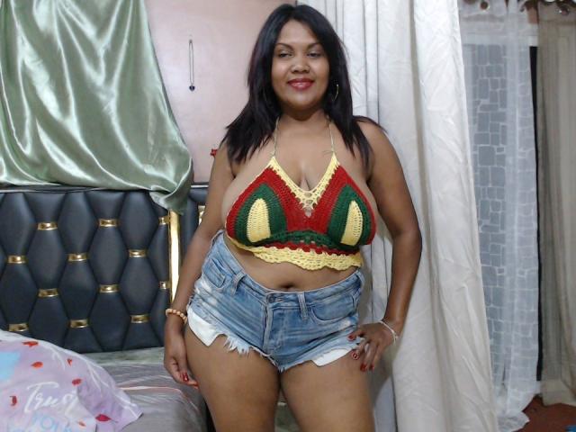 FelyLove has medium brown skin, black hair, and is smiling with red lipstick. She's wearing a green, yellow, and red crochet top with large boobs and ripped denim shorts.
