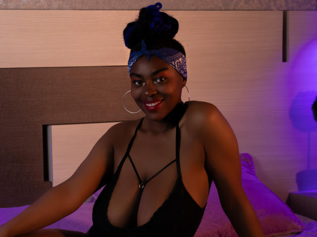 KrisMond with dark skin, blue hair bun, bandana, large hoop earrings, black halter top showing cleavage, smiling, sitting on bed, purple and orange lighting.
