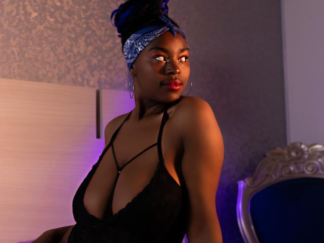 KrisMond, dark-skinned, wearing black lace lingerie, showing big tits, with blue headwrap, looking to the side, purple and pink light on her, sitting on ornate blue chair.