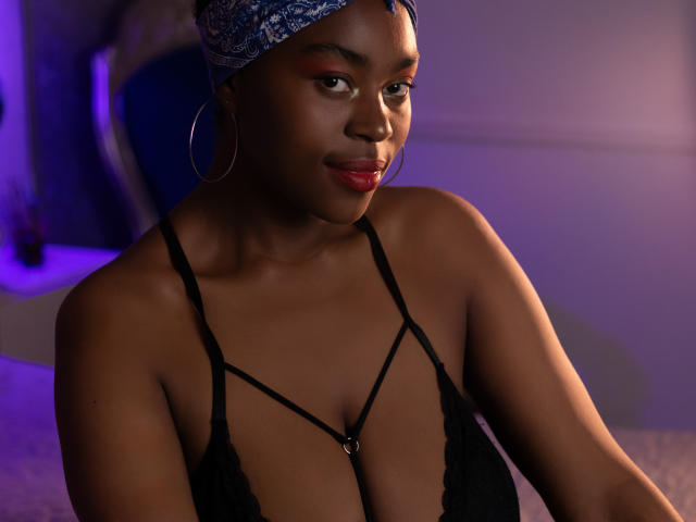 KrisMond with dark skin, wearing a black lace bra, hoop earrings, and a blue bandana, smiling with red lipstick, in soft purple lighting, showing her tits.