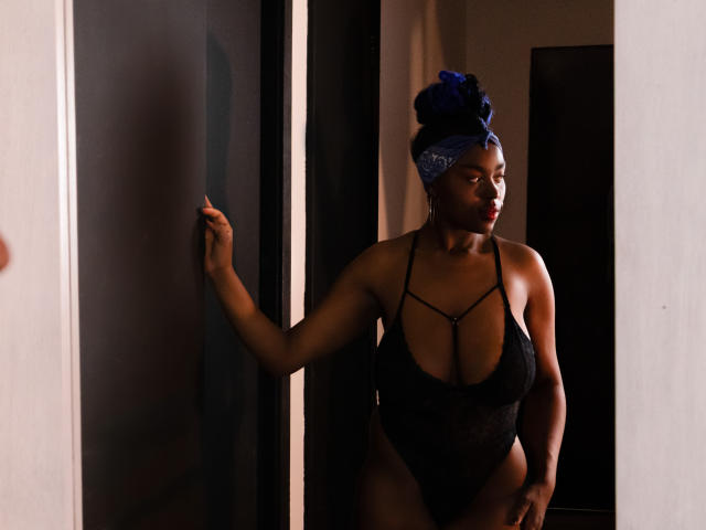 KrisMond stands in a dark doorway, wearing a black sheer lingerie set with strappy details, showing her big tits and curvy ass. She has blue hair in a bun.