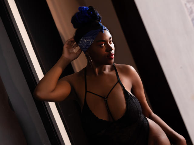 KrisMond with dark skin, blue headwrap, and red lipstick, wearing a black lace halter top that shows her big tits and slim waist, leaning against a dark wall.