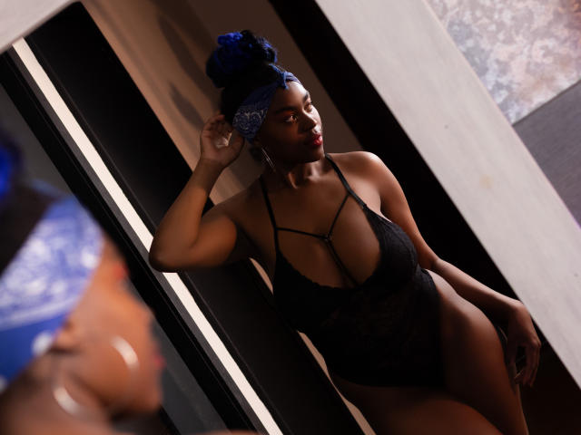 KrisMond with dark skin and blue bun, wearing black lace lingerie, standing in front of a mirror, showing her tits and ass, looking to the side with one hand up.