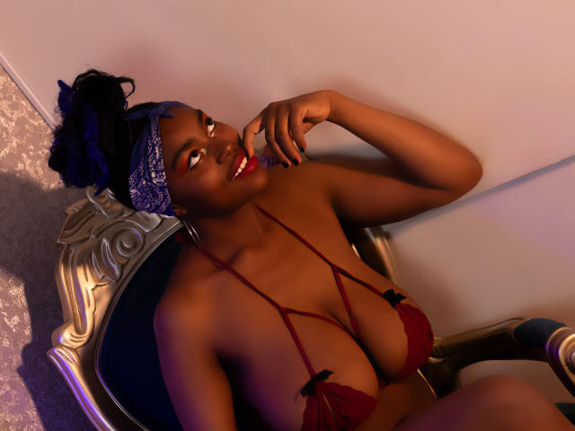 KrisMond with dark skin, blue hair, wearing red lace bra with black bows, smiling, finger touching lips, sitting on ornate chair, glowing light, sexy, curvy, large tits.