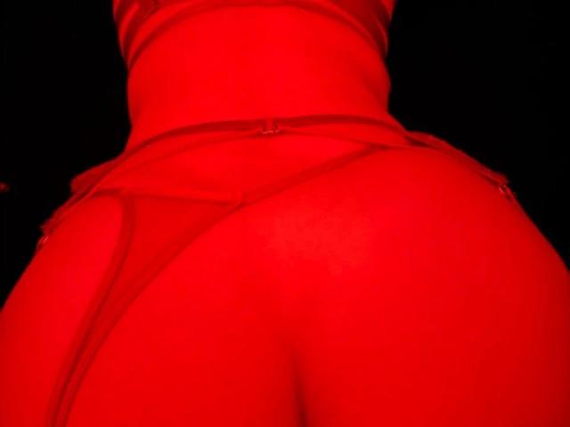 VaniXX-hot's red-covered ass, showing a tight, shiny, red garment with visible seams and a small zipper at the waist, photographed from a low angle against a dark background.