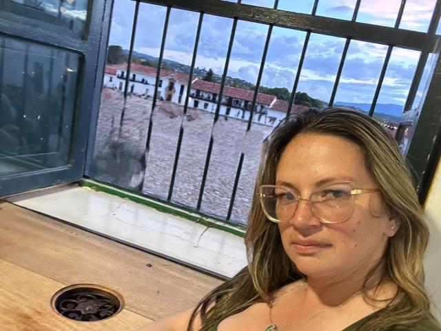 LadyLure with brown hair, glasses, and a black top, sitting at a table with a window showing a beach and white buildings outside. Her pussy is not visible.