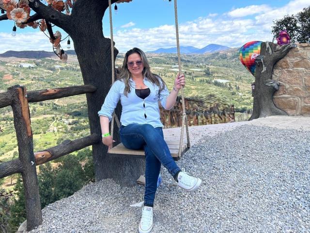 LadyLure with tits visible in a blue shirt, jeans, and white sneakers, sitting on a tree swing, holding a rope, smiling, wearing sunglasses, with a mountain view.