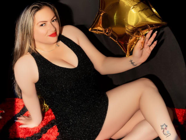 LadyLure, avec ses cheveux blonds, son rouge à lèvres rouge et ses tatouages, portant une robe noire scintillante, tenant un ballon en forme d'étoile dorée, assise sur une surface texturée rouge et noire..