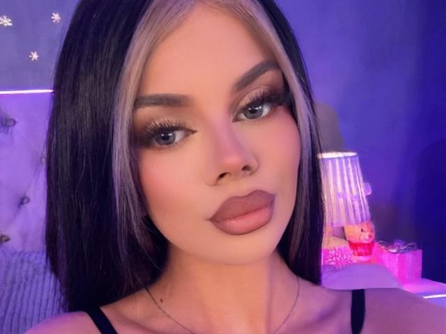 IvyRaven has long black and blonde hair, full eyebrows, long eyelashes, and puffy brown lips. She's wearing a black top with a thin necklace.