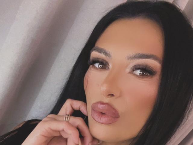 KinkyAmelie with full lips, dark eyes, and thick eyebrows, holding her chin with a ringed finger, has long black hair and a sexy, sultry look.