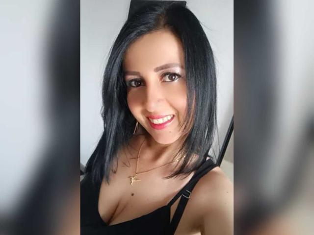 Briananice with black hair, smiling, wearing a black top, showing small tits, and a gold cross necklace, has red lipstick and brown eyes, standing in a selfie.