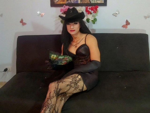 Sofia69xx with black cowboy hat, red lipstick, black lace dress, and gloves, holding bouquet, sitting on black couch, wearing fishnet stockings with lace pattern, showing tits.