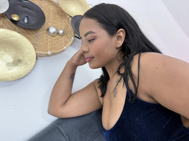 SilverLuna with tan skin and black hair, wearing a blue velvet dress, resting her chin on her hand, showing her small tattoo and looking down. Boobs visible.