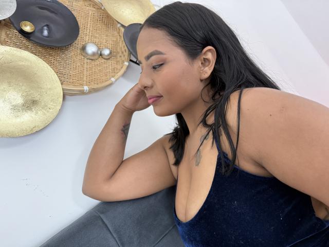 SilverLuna with black hair, medium skin, wearing a deep blue velvet dress with a low-cut neckline, showing her tits, arm tattoo, and pink lipstick, resting her chin on her hand.