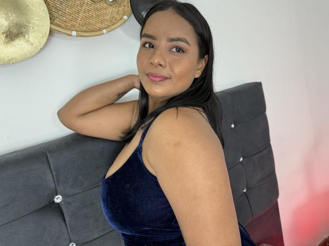 SilverLuna, with brown skin and long black hair, wearing a dark blue velvet dress, showing her tits, smiling, arm raised, standing in front of a grey tufted headboard.