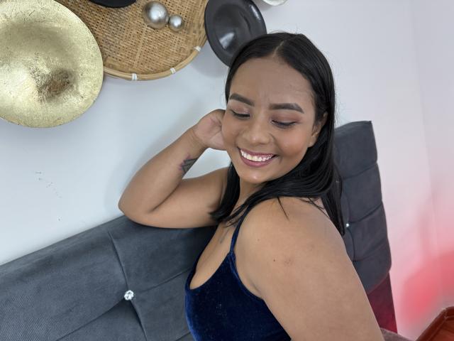 SilverLuna with black hair, smiling, wearing a blue velvet sleeveless top, showing a tattoo on her arm, sitting on a grey tufted sofa, hand near her cheek.
