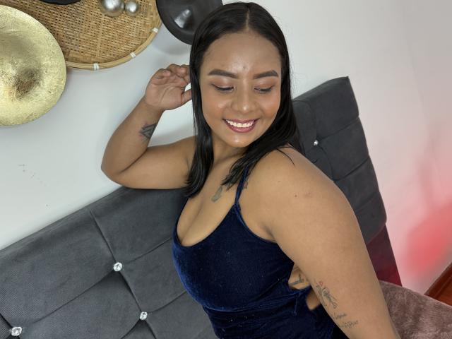 SilverLuna with black hair, smiling, wearing a dark blue velvet dress, showing her tits and tattoos on her arm and side, standing in front of a gray tufted headboard.