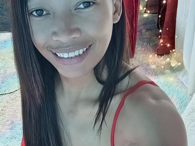 RedBellah with long brown hair, smiling, wearing a red spaghetti strap top, showing her teeth and white boobs in a selfie. Fairy lights in the background.
