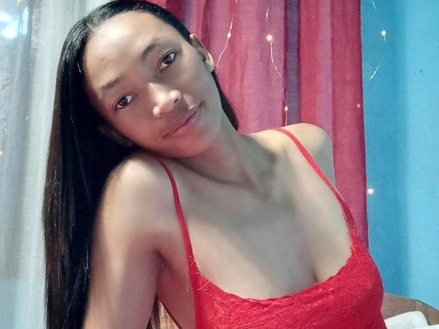 RedBellah with long black hair, smiling slightly, wearing a red lace spaghetti top, showing her small tits and slim collarbone, against a blue and red curtain.