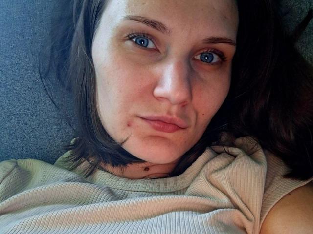 ErrosAy has blue eyes, light skin, and brown shoulder-length hair, wearing a ribbed shirt, with a small pimple on her chin, lying on a gray couch.
