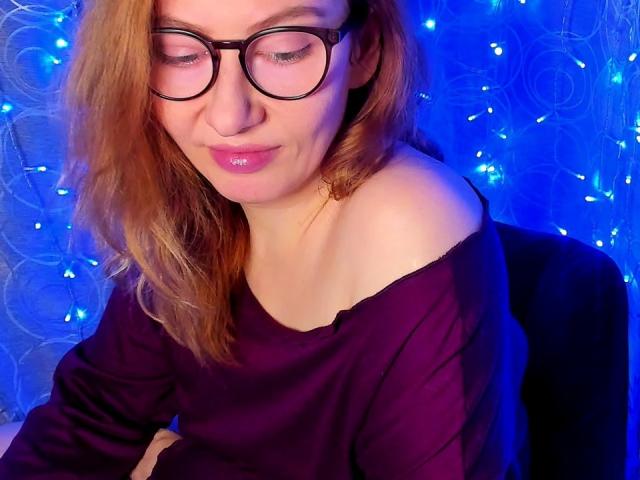 PaulinaSunny with glasses, pink lips, and wavy red hair, wearing an off-shoulder purple top, sitting with a slight smile, blue fairy lights in the background.