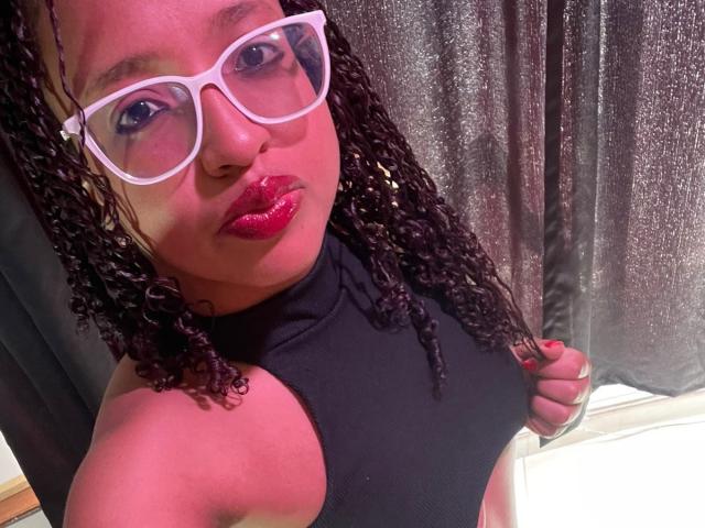 ArtemisaS with curly black hair, red lipstick, and white glasses, wearing a black halter top, showing her bare shoulders and midriff, holding the top.