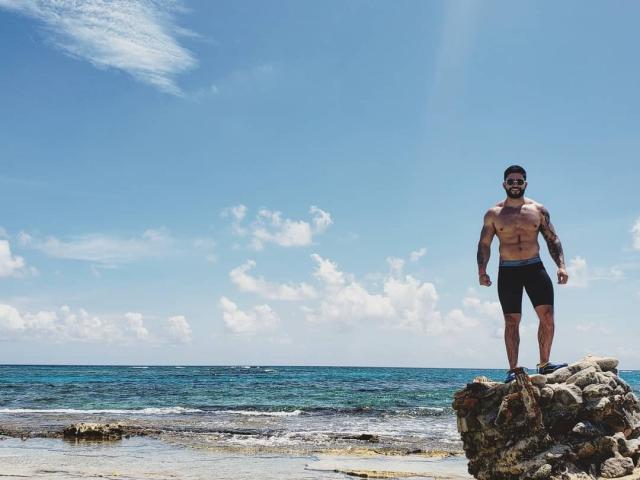 Lukas stands on a rocky outcrop by the ocean, shirtless, wearing black shorts. He has a muscular build, short dark hair, and a beard. His tattooed arms are slightly flexed. The clear blue sky with scattered white clouds contrasts with the turquoise sea. Lukas's right foot is on a larger rock, his left on a smaller one. Sunlight from the top right highlights his upper body. The beach and sea are in the background. Image has a realistic, natural style.